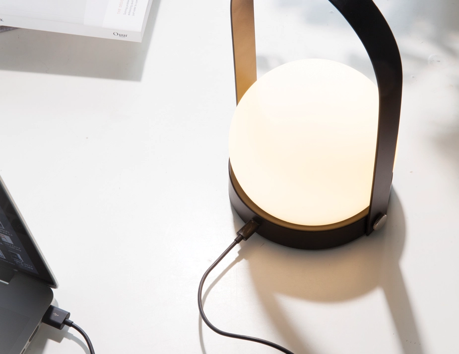 Close-up of a glowing black Carrie Table Lamp connected by a charging cable to a laptop