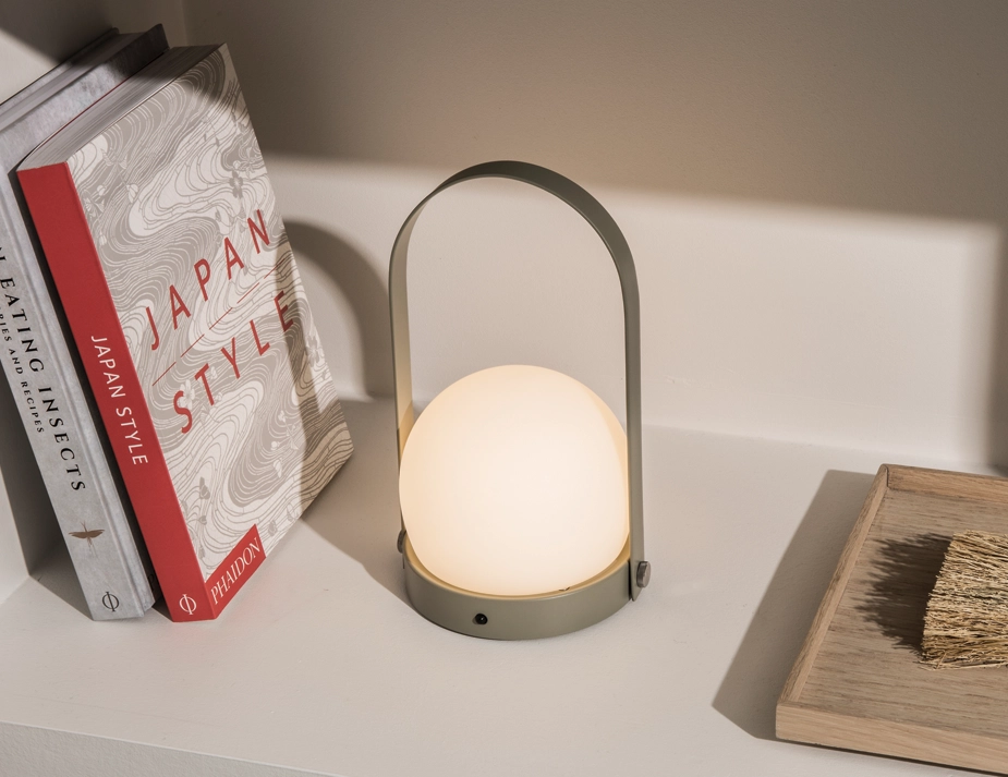 Glowing olive green Carrie Table Lamp beside books and a wooden tray on a white shelf