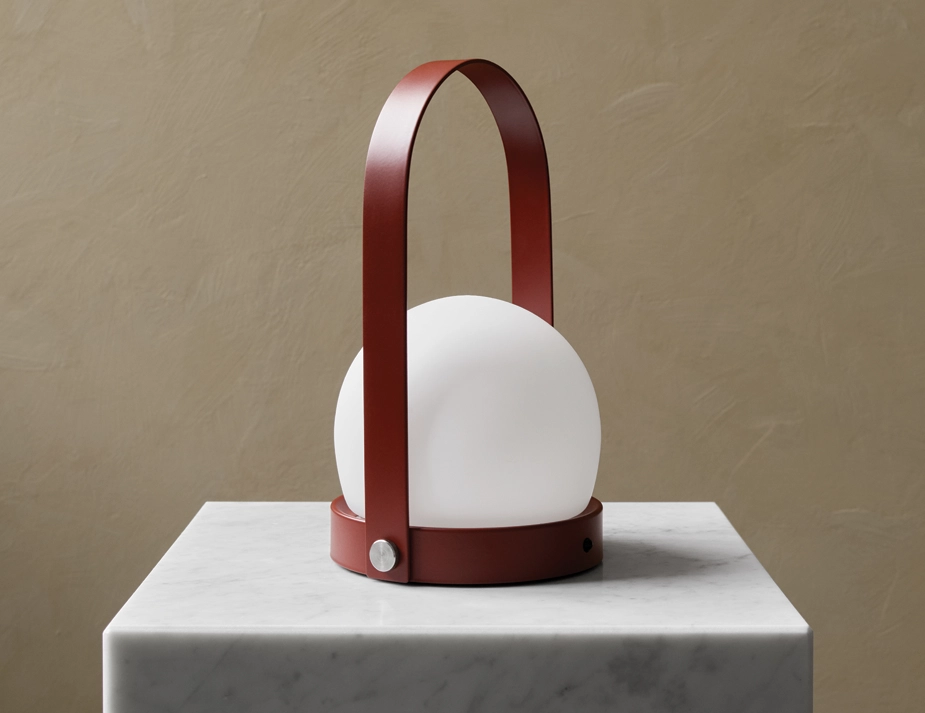 Burned red Carrie Table Lamp with a white globe shade and arched handle on a pale square plinth